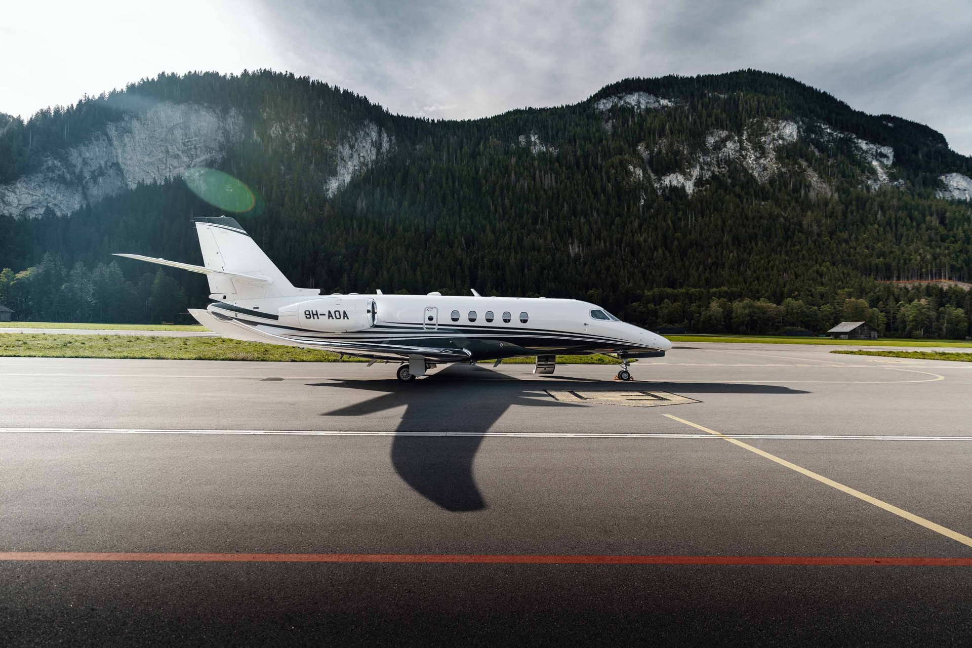 et privé Cessna Citation Latitude d’Albinati Aeronautics stationné sur le tarmac d’un aéroport alpin, avec en arrière-plan des montagnes et une forêt sous un ciel partiellement nuageux.